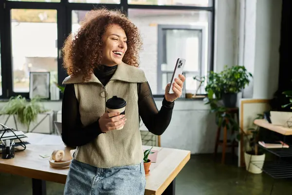 A cheerful woman with curly hair is smiling as she looks at her phone while holding a coffee cup. — Stock Photo