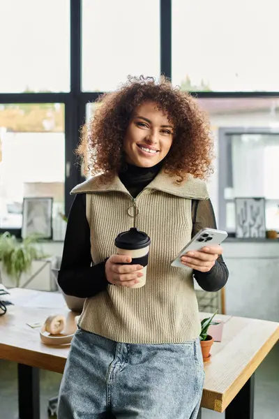 La jeune femme aux cheveux bouclés sourit tout en tenant une tasse de café et un téléphone au bureau — Photo de stock