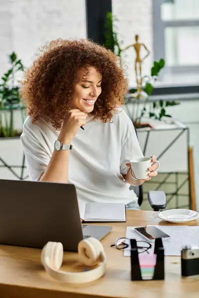 Une femme aux cheveux bouclés sirote du thé, immergée dans son espace de travail créatif, souriant joyeusement. — Photo de stock