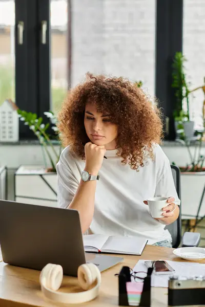 Una mujer con el pelo rizado se sienta en un escritorio, profundamente comprometida con su trabajo mientras bebe café. - foto de stock