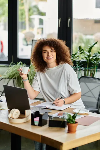 A cheerful woman holds a cup of coffee while writing in a cozy workspace with plants. — Stock Photo