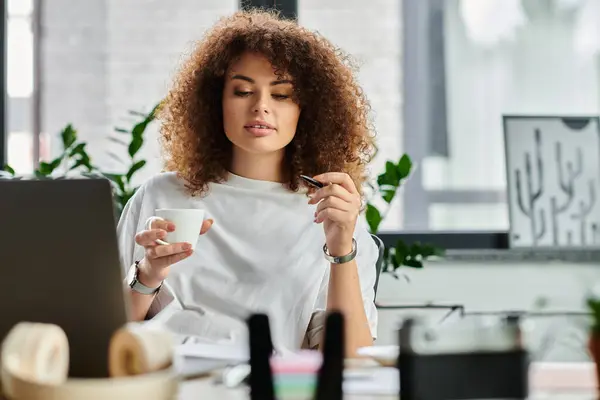 A woman with curly hair takes a moment to sip coffee in a modern workspace filled with greenery. — Stock Photo