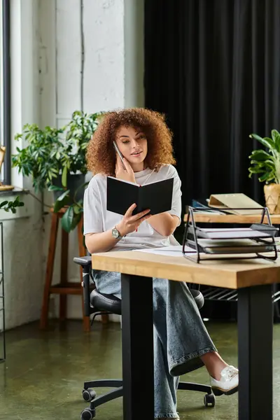 Curly haired woman sits thoughtfully while reading a notebook in a stylish office setting. — Stock Photo