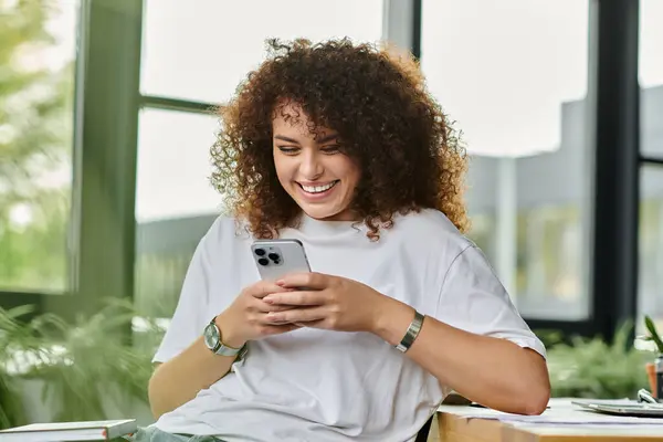 A joyful young woman smiles as she engages with her smartphone in a sunny office environment. — Stock Photo