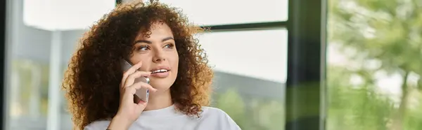 A woman with curly hair engages in a thoughtful discussion on her smartphone in a sunlit room. — Stock Photo