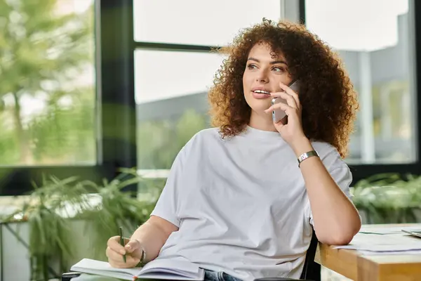 A woman with curly hair engages in a lively discussion on her mobile while taking notes. — Stock Photo