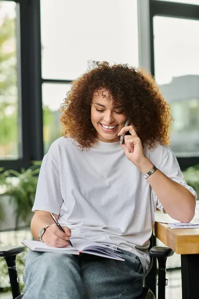 Una donna sorride e scrive appunti durante una vivace conversazione telefonica in una stanza luminosa. — Foto stock