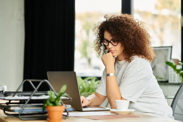 A woman with curly hair reflects while focusing on her laptop in a vibrant office setting. — Stock Photo