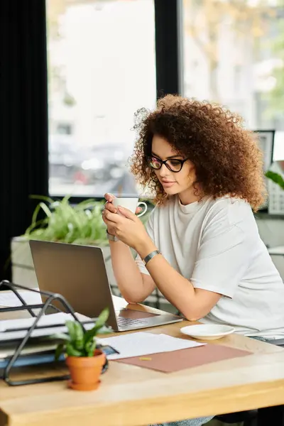 A woman sits at a wooden table, savoring her coffee as she focuses on her laptop. — Stock Photo