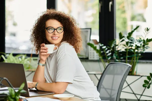 Eine Frau mit lockigem Haar nippt an Tee und konzentriert sich auf ihre Arbeit in einem gemütlichen Büroraum. — Stockfoto