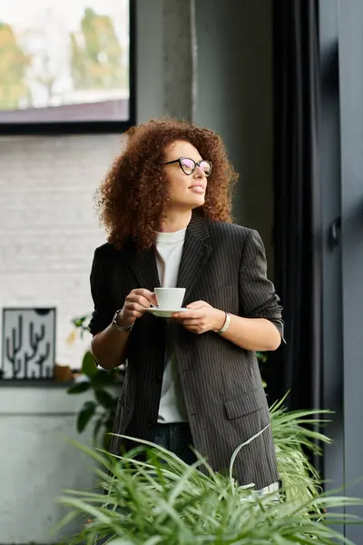 A woman with curly hair sips coffee and admires the view from a stylish interior room. — Stock Photo