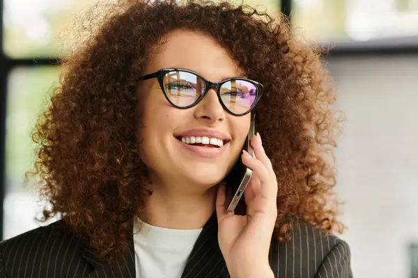 A smiling woman with curly hair engages in a lively conversation on her phone at work. — Stock Photo