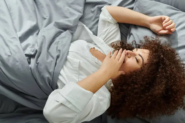 La mujer yace cómodamente en su cama, estirándose y bostezando mientras la luz de la mañana ilumina la habitación. - foto de stock