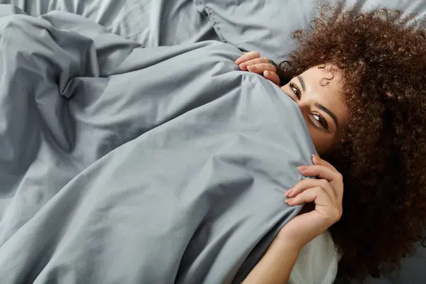 A woman hides playfully under a blanket, revealing a joyful expression while relaxing in bed. — Stock Photo