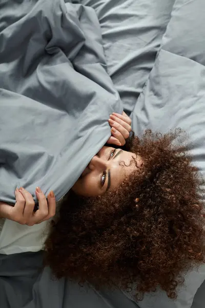 A woman peeks out from beneath gray blankets, enjoying a playful moment in bed. — Stock Photo
