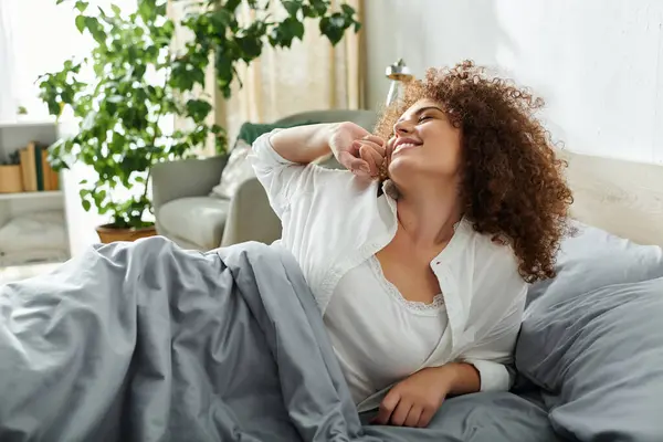 Una mujer se estira alegremente en su acogedor dormitorio, disfrutando de la tranquila luz de la mañana. - foto de stock