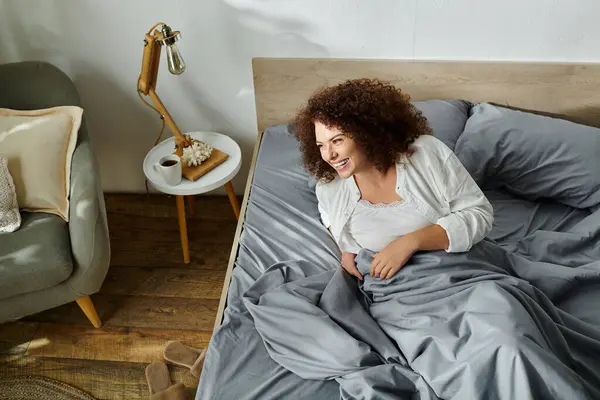 A smiling woman lies comfortably in bed, surrounded by soft blankets, enjoying the morning light. — Stock Photo