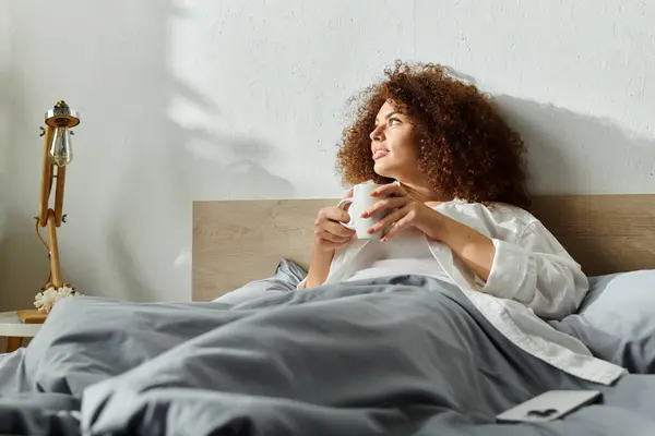 A thoughtful woman sits in bed, sipping a warm beverage and contemplating her morning. — Stock Photo