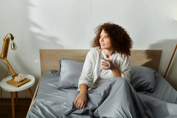 A woman sits on her bed, savoring a warm drink while embracing the calm of the morning. — Stock Photo