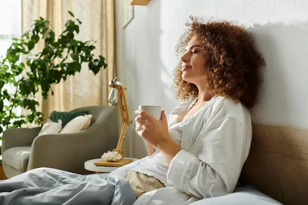 Una mujer relajada saborea su té, abrazando la tranquilidad en un dormitorio soleado lleno de vegetación. - foto de stock