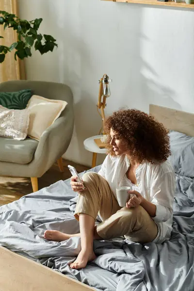 The woman sits comfortably on her bed, enjoying a warm cup of coffee while using her phone. — Stock Photo