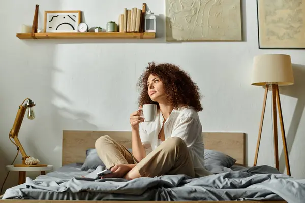 A woman sits cross legged on the bed, contemplating with a cup of tea in her hand. — Stock Photo