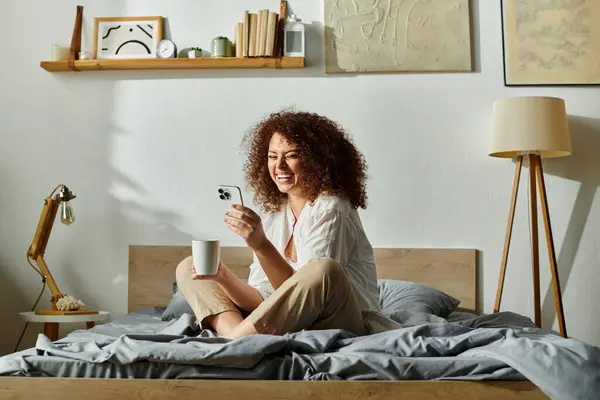 Una mujer alegre disfruta de su café de la mañana mientras se conecta con su teléfono en un dormitorio luminoso. - foto de stock