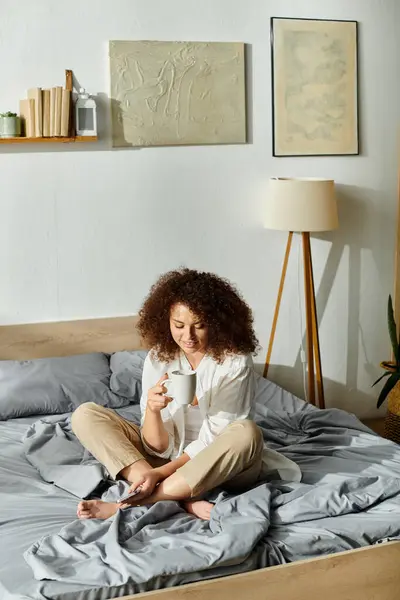 A woman sits cross legged on a bed holding a cup, embracing a tranquil morning vibe. — Stock Photo