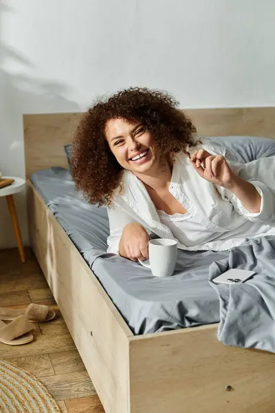 The woman lounges on her bed, smiling joyfully as she holds a coffee cup. — Foto stock