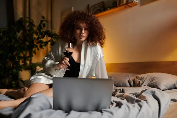 A woman enjoys her evening on the bed, sipping wine while using her laptop. — Stock Photo