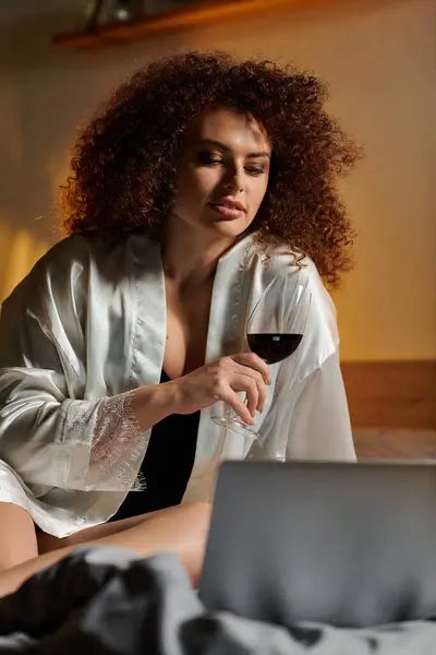 Una donna elegante si rilassa a letto, sorseggiando vino e coinvolgendo con il suo computer portatile in un'atmosfera calda. — Foto stock