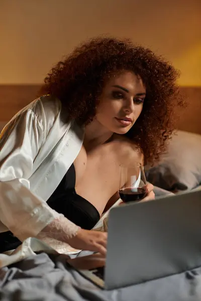 A woman with curly hair wears a satin robe, sipping wine as she focuses on her laptop. — Stock Photo