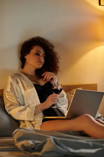 Entspannt in einem ruhigen Schlafzimmer schlürft eine Frau Wein, während sie mit ihrem Laptop beschäftigt ist. — Stockfoto