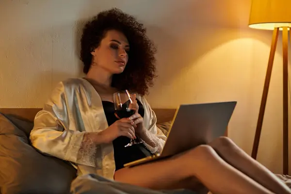 A woman enjoys a glass of red wine while focused on her laptop at night. — Stock Photo