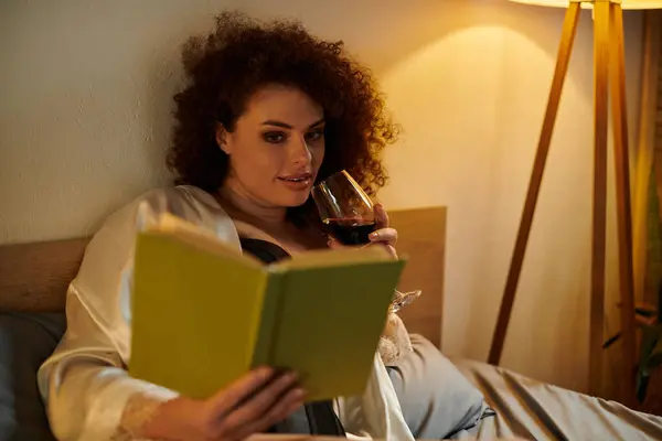 Détente dans un espace cosy, une femme lit un livre tout en savourant un verre de vin, rayonnant de confort. — Photo de stock