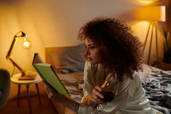 Dans une chambre sereine, une femme immergée dans son livre savoure une soirée avec un verre de vin. — Photo de stock