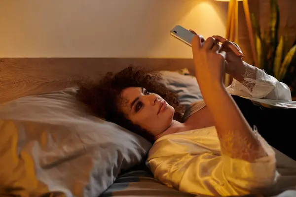 A woman lounges comfortably in bed, engaged with her smartphone amidst soft lighting. — Stock Photo