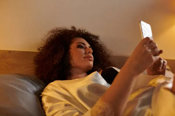 A woman reclines comfortably in bed, engrossed in her smartphone, surrounded by a warm atmosphere. — Stock Photo