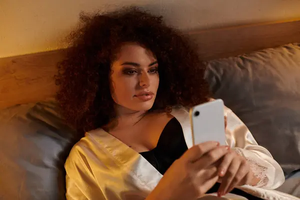 A woman with curly hair enjoys time in bed, absorbed in her smartphone under warm lighting. — Stock Photo