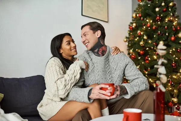 A couple shares smiles and warmth while sipping drinks beside a beautifully decorated tree. — Stock Photo