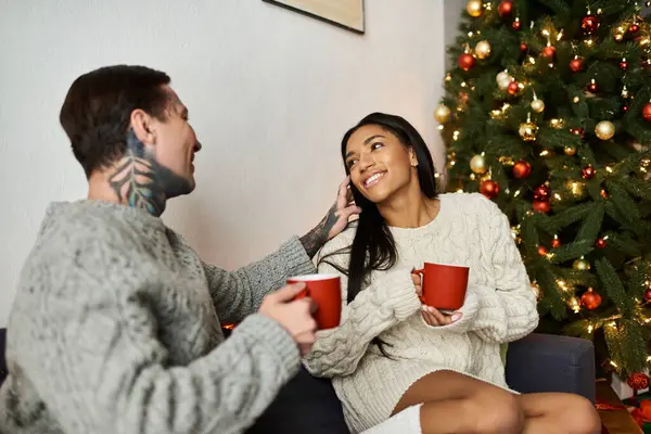 Una pareja disfruta de su tiempo juntos, riendo y bebiendo bebidas calientes cerca de un árbol de Navidad festivo. - foto de stock