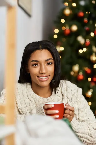 Una mujer alegre se relaja, tomando una bebida caliente frente a un árbol bellamente decorado. - foto de stock