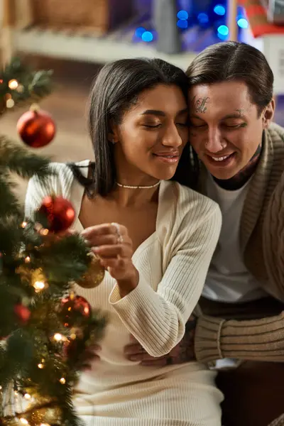 A couple embraces each other while adorning their Christmas tree with ornaments in their warm home. — Stock Photo