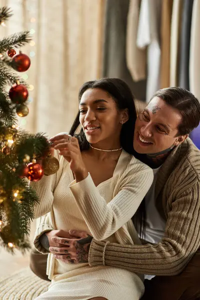 Together they hang ornaments on their beautifully lit Christmas tree, sharing laughter. — Stock Photo