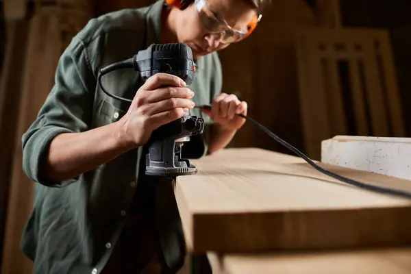 A dedicated carpenter is focused on shaping wood using a power tool in her workshop, showcasing her craftsmanship. — Stock Photo