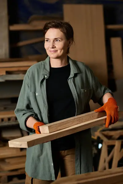 Eine talentierte Schreinerin lächelt, während sie Holzbohlen trägt, eingetaucht in ihr Handwerk in einer geschäftigen Werkstatt. — Stockfoto