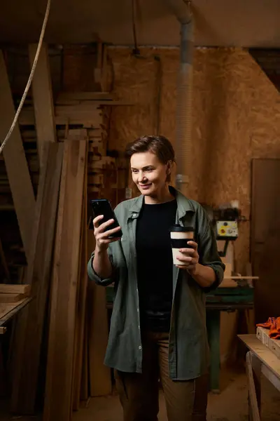 In a rustic workshop, a female carpenter checks her phone while balancing a coffee cup in hand. — Stock Photo