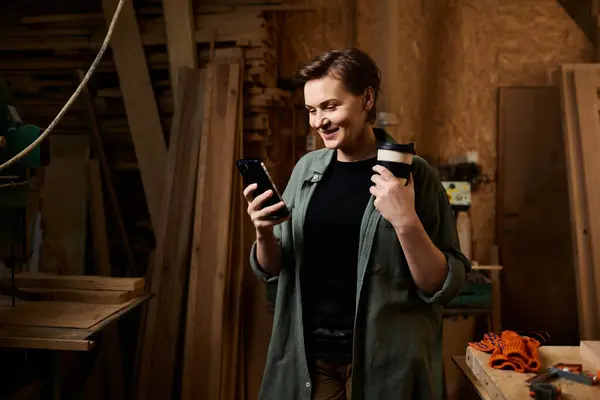 A talented carpenter takes a moment to relax, holding a coffee cup while looking at her phone in the workshop. — Stock Photo