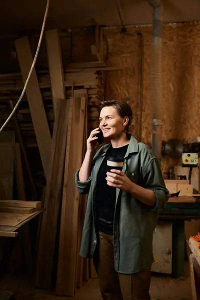 A skilled female carpenter chats on the phone, sipping coffee in her busy wood workshop filled with materials. — Stock Photo