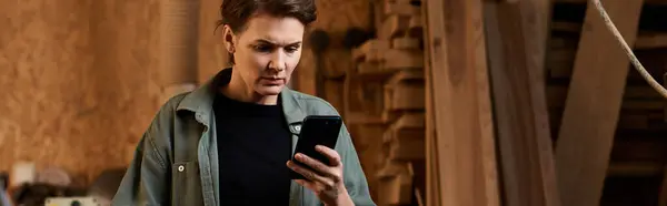 Талантливая женщина-плотница рассматривает важную информацию на своем телефоне среди инструментов и проектов по деревообработке. — стоковое фото